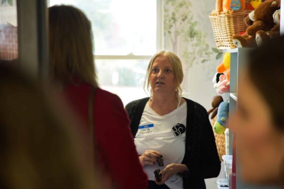 A woman speaking to another woman inside a building with stuffed animals in the background.