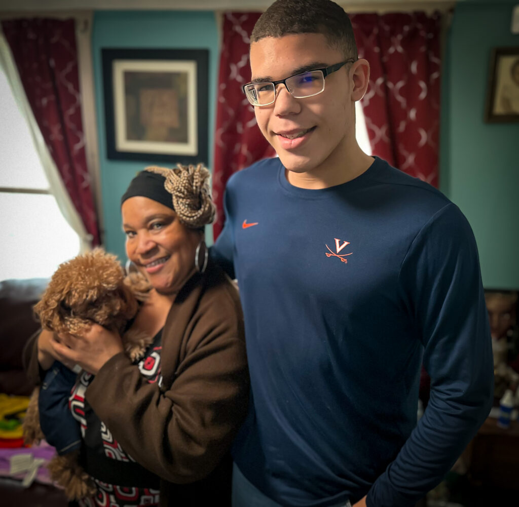 A grandma and grandchild posing for a picture inside of a house. The grandma is holding onto a dog.